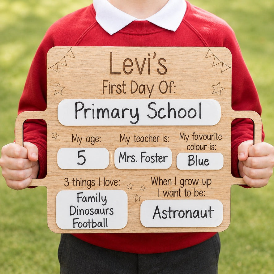 Personalised wooden first day of school sign with dry wipe panels, held by a child wearing a red school jumper outdoors. Features custom name, school year, age, teacher, favourite colour, favourite things and future career, handcrafted from oak veneer with laser engraving.